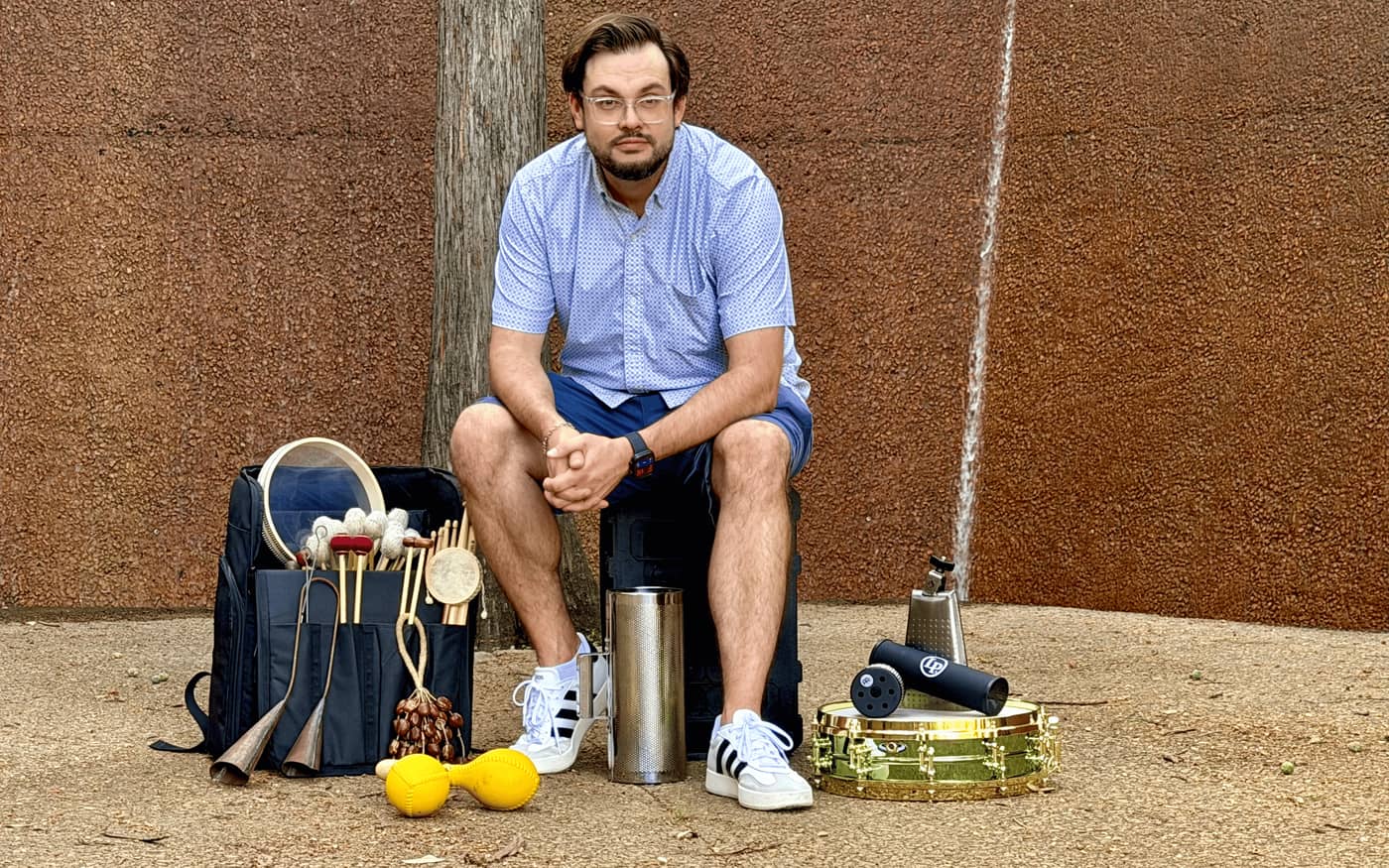 Taylor Dwyer seated against a rust-red wall with his travel kit laid out — a loaded mallet bag, frame drum, maracas, cowbell, and brass piccolo snare